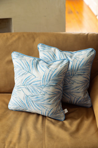 Two Harbor Palmetto Pillows, featuring a blue botanical pattern and blue piping, are placed on a tan leather couch. The beige wall and wooden floor in the background enhance the coastal aesthetic of the pillows.