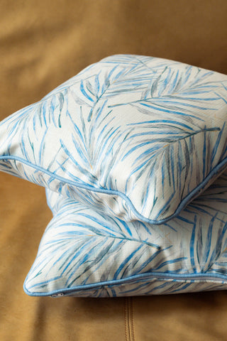Two Harbor Palmetto Pillows featuring a blue palm leaf botanical pattern and coastal design rest on a tan fabric surface. Made in the USA, the pillows are stacked one atop the other, highlighting their soft texture and vibrant tropical style.