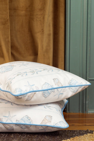 Two Sullivan’s Shore Pillows, featuring white fabric adorned with light blue seashell and starfish patterns, are stacked on a wooden floor beside a green door and a brown curtain, bringing charming seaside decor into the room.