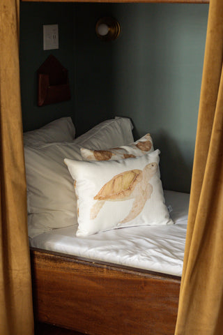 A cozy bed with white linens and pillows, including the decorative Tidal Travelers Prefilled Lumbar Pillow, is framed by mustard yellow curtains in a softly lit room.