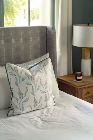 A neatly made bed features the elegant Shoreline Vines Pillow, placed beside a wooden nightstand adorned with a lamp and candle. Soft natural light filters through white curtains by the window, creating an inviting atmosphere that showcases the organic charm of your decor.