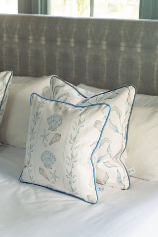 Two decorative pillows, including the Sullivan’s Shore Pillow featuring seashell and seaweed patterns, rest on a neatly made bed with a gray upholstered headboard and crisp white sheets. Sunlight filters through the window in the background, enhancing the relaxing vibe of this seaside home decor.