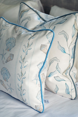 Two decorative Shoreline Vines Pillows with blue piping rest on a white bed. Featuring sea-inspired designs—seashells, starfish, and seaweed in soft blue and beige tones—these organic pillow covers bring a tranquil coastal touch to any bedroom. Add seaside charm and elegance to your space with the Shoreline Vines Pillow collection.