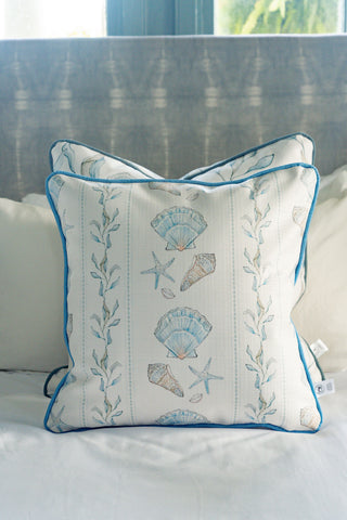 Two Sullivan’s Shore Pillows with blue piping and a seashell, starfish, and coral pattern rest against a white bed and gray upholstered headboard, bringing a touch of seaside home decor to the space as light streams in from a window in the background.