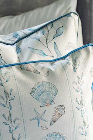 Two Sullivan’s Shore Pillows featuring a seaside decor design with seashells, starfish, and seaweed in blue and beige tones. These decorative coastal pillows have blue piping along the edges and are displayed on a bed, making them perfect for adding a charming touch to any cottage pillow collection.