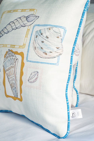 The Coastal Curio Pillow is a square coastal pillow with blue trim, featuring seashell illustrations framed in yellow, blue, and beige on the cover. Perfect for adding a touch of beach home decor to your space, it looks beautiful resting on a white bed.