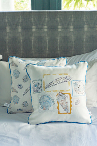 Two Coastal Curio Pillows with charming seashell prints rest against each other on a white bed, complemented by a gray upholstered headboard and a window in the background—perfect for adding a touch of coastal home decor to your space.