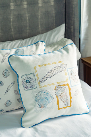 Two Coastal Curio Pillows with blue piping rest on a bed. The front pillow showcases a collection of seashell illustrations, while the back pillow features a repeating seashell pattern. Perfect for coastal home decor, the headboard and white bedding are partially visible.