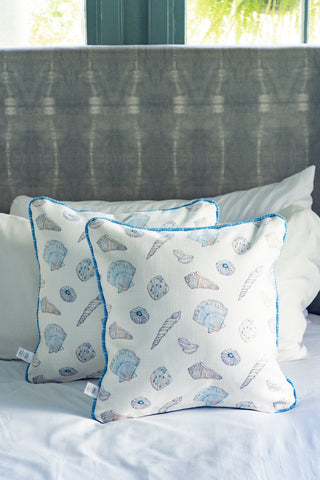 Two Coastal Curio Pillows featuring blue and beige seashell patterns rest on a white bed, set against a grey upholstered headboard with a window showcasing green scenery in the background.