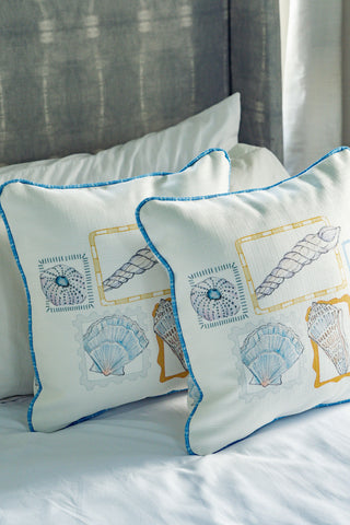 Two Coastal Curio Pillows featuring blue and yellow seashell and sea-themed illustrations rest against a white pillow and gray headboard on a bed, adding the perfect touch of coastal home decor.