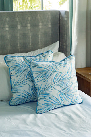 Two Harbor Palmetto Pillows, featuring white fabric with blue leaf patterns, rest against a grey upholstered headboard on a neatly made bed, adding a subtle botanical flair to the room. The tranquil setting is enhanced by a wooden nightstand and softly draped curtains at the window, creating an inviting coastal atmosphere.