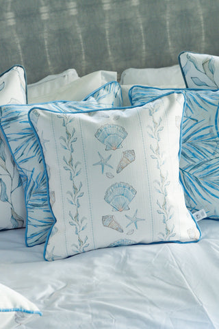 The Sullivan’s Shore Pillow, with its blue and white patterns featuring seashells, starfish, and seaweed, creates charming seaside decor when arranged on a bed with a light gray headboard in the background.