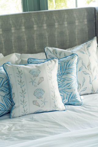 Four Sullivan’s Shore Pillows, featuring blue and beige seashell and leaf patterns, are arranged on a neatly made bed with a light grey upholstered headboard in the background, adding a charming touch of cottage decor to the room.