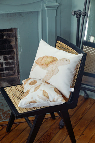 The Tidal Travelers Prefilled Lumbar Pillow, featuring charming turtle illustrations including a sea turtle design, sits on a black wooden chair with a woven rattan seat next to a fireplace and wooden floor, adding a touch of coastal decor to the space.