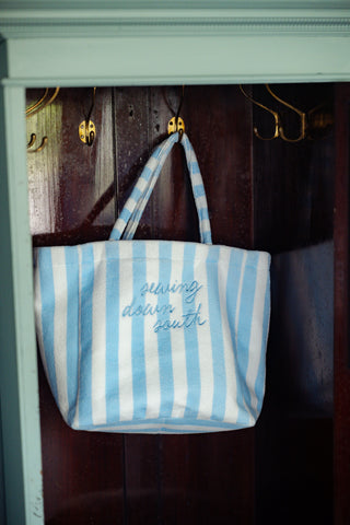 A Weekend Getaway Stripe Terry Tote, featuring classic blue and white stripes and “sewing down south” embroidery, hangs on a hook inside a wooden closet—ready to accompany you on your next beach adventure or weekend escape.