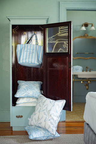 An open wooden cabinet displays stacks of blue and white patterned pillows spilling onto the floor, while the Weekend Getaway Stripe Terry Tote in blue stripes sits nearby. In the background, a vintage sink and mirror enhance the relaxed atmosphere, making this space perfect for a weekend escape.