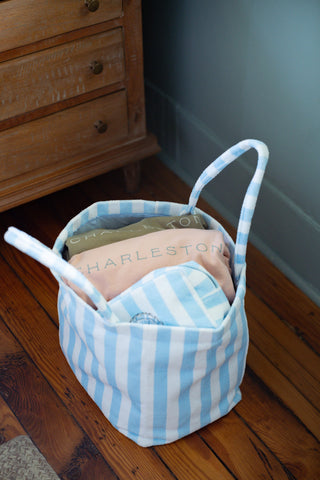 A Weekend Getaway Stripe Terry Tote, in blue and white stripes and reminiscent of a classic beach bag, sits on a wooden floor holding folded blankets and pillows—including one labeled CHARLESTON—next to a wooden dresser.