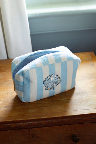 The Weekend Getaway Striped Terry Cosmetic Bag, featuring blue and white stripes and a shell design, rests on a wooden surface by a window with white curtains—an essential piece from the Shore capsule collection, ideal for your next weekend escape.