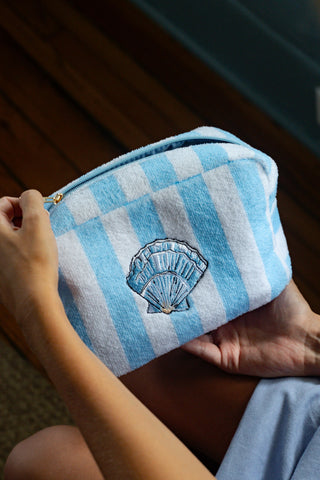 A person holds the Weekend Getaway Striped Terry Cosmetic Bag, featuring blue and white stripes and a blue seashell motif. This zippered pouch, crafted from soft terry material as part of the Shore Capsule Collection, is ideal for a weekend getaway.
