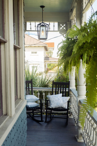 Two black rocking chairs with patterned Outdoor Washed Ashore Pillows sit on a cozy porch with white railings, hanging ferns, and a lantern above. Coastal decor accents the space, while a house and greenery are visible in the background.