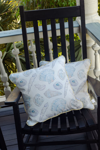 A black wooden rocking chair with two Outdoor Washed Ashore Pillows—white cushions featuring light blue seashell patterns—sits on a porch with a white railing and greenery in the background, adding an inviting touch of coastal decor.