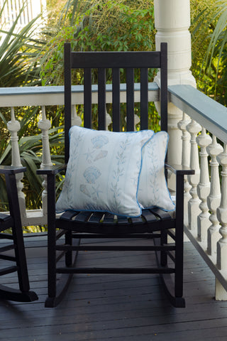 A black wooden rocking chair with an Outdoor Sullivan’s Shore Pillow in light blue sits on a porch with white railings and greenery in the background.