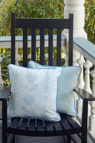 A black wooden chair on a porch holds two light blue Outdoor Sullivan’s Shore Pillows, one embroidered with seashells and starfish. Lush green foliage and white railing surround the setting.