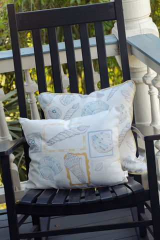 A black wooden rocking chair sits on a porch with a white railing and greenery in the background, adorned with an outdoor pillow and the Outdoor Coastal Curio Lumbar Pillow, both featuring seashell designs.