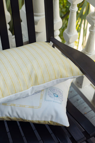 Two patterned pillows—an Outdoor Coastal Curio Lumbar Pillow in yellow stripes and a geometric lumbar pillow—rest on a black slatted outdoor bench. White railing and green foliage are visible in the background.