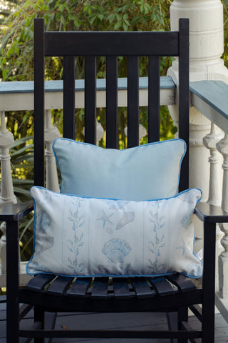 A black wooden rocking chair sits on a porch with two light blue pillows—one solid and the other, the Outdoor Sullivan’s Shore Extended Lumbar Pillow, showcasing a charming seashell and starfish design—while lush greenery provides a tranquil backdrop.