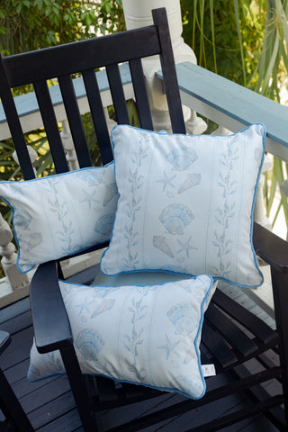 An Outdoor Sullivan’s Shore Extended Lumbar Pillow with light blue hues and seashell and starfish patterns rests on a black wooden rocking chair on a porch, surrounded by greenery and a white railing.