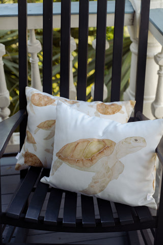 Two Outdoor Tidal Travelers Lumbar Pillows featuring turtle illustrations rest on a black slatted chair on a porch, with green foliage and a white railing in the background.