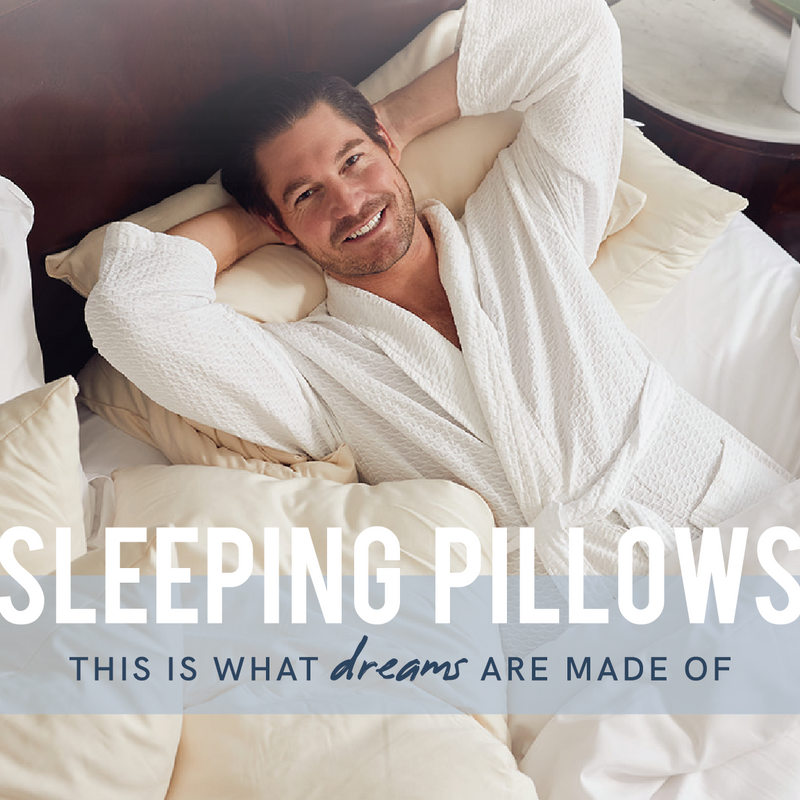 A smiling man in a white bathrobe lies on a bed surrounded by plush pillows. Text overlay reads: SLEEPING PILLOWS - THIS IS WHAT dreams ARE MADE OF.