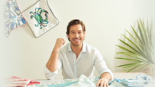 A man with brown hair and a light shirt sits at a table covered with colorful fabrics. He smiles at the camera, raising one fist. Behind him, a green plant and decorative cloths hang on a white wall.
