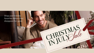 Promotional banner featuring a smiling man in a beige knit sweater seated indoors, with large text reading 'Christmas in July Sale' on a tilted white sign and red ribbon graphics across the frame.
