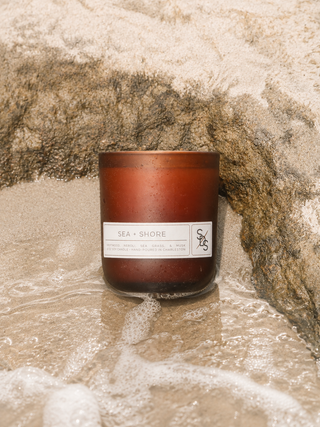 A brown Sea + Shore Limited Edition Candle rests on wet sand between rocks, as waves gently wash around its base. Hand-poured in Charleston, this candle features a fresh coastal scent that captures the spirit of the ocean.