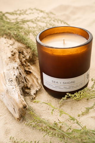 The Sea + Shore Limited Edition Candle, housed in brown glass, rests on sand alongside driftwood and green sprigs, evoking a coastal, natural aesthetic. Hand-poured in Charleston, this candle fills your space with a refreshing coastal fragrance inspired by the serene ambiance of the shore.