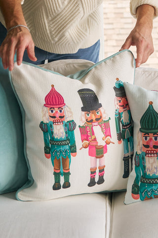 A person places the Pastel Nutcracker Pillow, featuring colorful nutcracker designs, on a light couch. Theyre wearing a cream sweater, with a window and blurred outdoor view in the background.