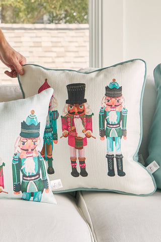 Someone places the Pastel Nutcracker Pillow—featuring colorful nutcracker illustrations in pastel uniforms—on a white couch, next to a smaller matching pillow for a festive holiday look.