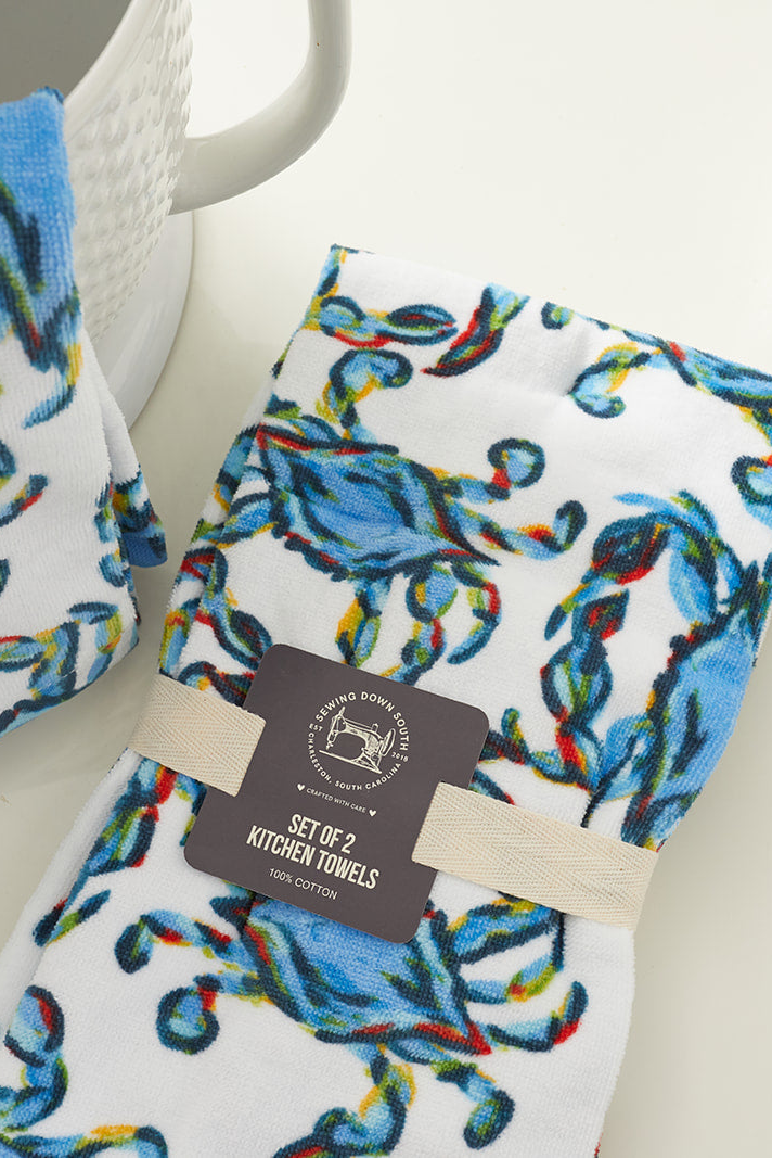 The Crab Craze Kitchen Towels, 2 pack, feature colorful blue and green crab prints. These oversized cotton towels add a coastal touch to your kitchen and come folded with a paper label and beige band near a white textured bowl.