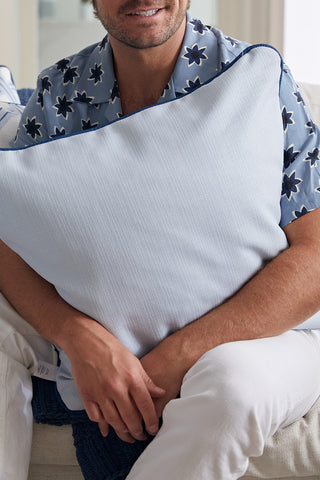 A man in a blue floral shirt and white pants sits on a sofa, holding the Oversized Mod Stella Tile Pillow in front of his chest. Only his lower face and hands are visible, drawing attention to the pillow’s bold, decorator style.
