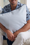 A person in a blue floral shirt and white pants sits on a couch, holding a light blue Mod Stella Tile Pillow from the Modern Collection. The pillows subtle geometric design enhances the relaxed, comfortable setting.