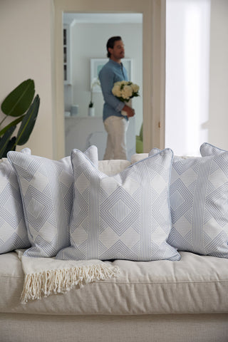 An Oversized Mod Geo Diamond Pillow and a light throw blanket are on a beige sofa. In the background, a man holding white flowers stands in the doorway, slightly out of focus.