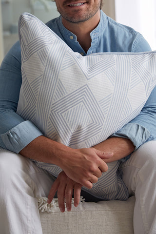 A man in a light blue shirt and white pants sits on a couch, smiling and holding the Oversized Mod Geo Diamond Pillow from Sewing Down South, which features a light blue and white geometric pattern.
