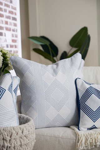 A beige sofa features modern pillows, including the Oversized Mod Geo Diamond Pillow in blue with a geometric pattern; smaller pillows in navy and white diamond designs, a green plant, and a window complete this Sewing Down South-inspired scene.