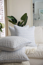 A cozy living room features a white sofa with four Mod Mini Crossing Tiles in Blue pillows. Two green plants add natural accents, complementing the Modern Collection decor. Sunlight streams through the window, enhancing the serene southern aesthetic.