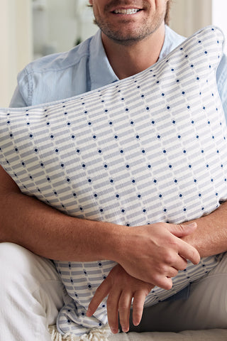 A man in a light blue shirt and white pants sits, smiling, as he hugs the Oversized Mod Mini Crossing Tiles in Blue Pillow—bringing modern style to Southern home decor.