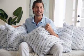 A smiling man in a light blue shirt and white pants sits on a sofa, holding the Oversized Mod Mini Crossing Tiles in Blue Pillow among matching pillows, with a large green plant and natural light accenting this inviting Southern home decor scene.