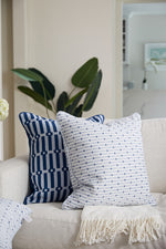 Two Mod Mini Crossing Tiles in Blue pillows from the Modern Collection on a light sofa, with a soft beige throw draped over its arm. A large leafy plant in the background adds greenery to this bright, neutral room.