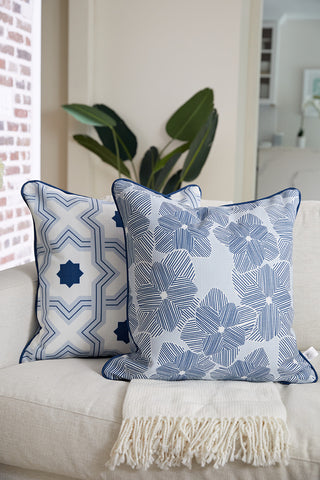 An Oversized Mod Flora Pillow from Sewing Down South with a blue floral pattern sits on a light sofa draped with a cream fringed throw; a leafy green plant is visible in the background.